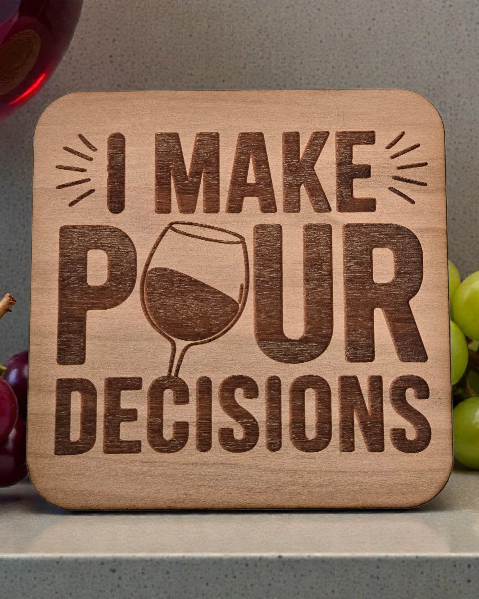 Wooden coaster with 'I Make Pour Decisions' text and a wine glass graphic, surrounded by grapes and a glass of red wine.