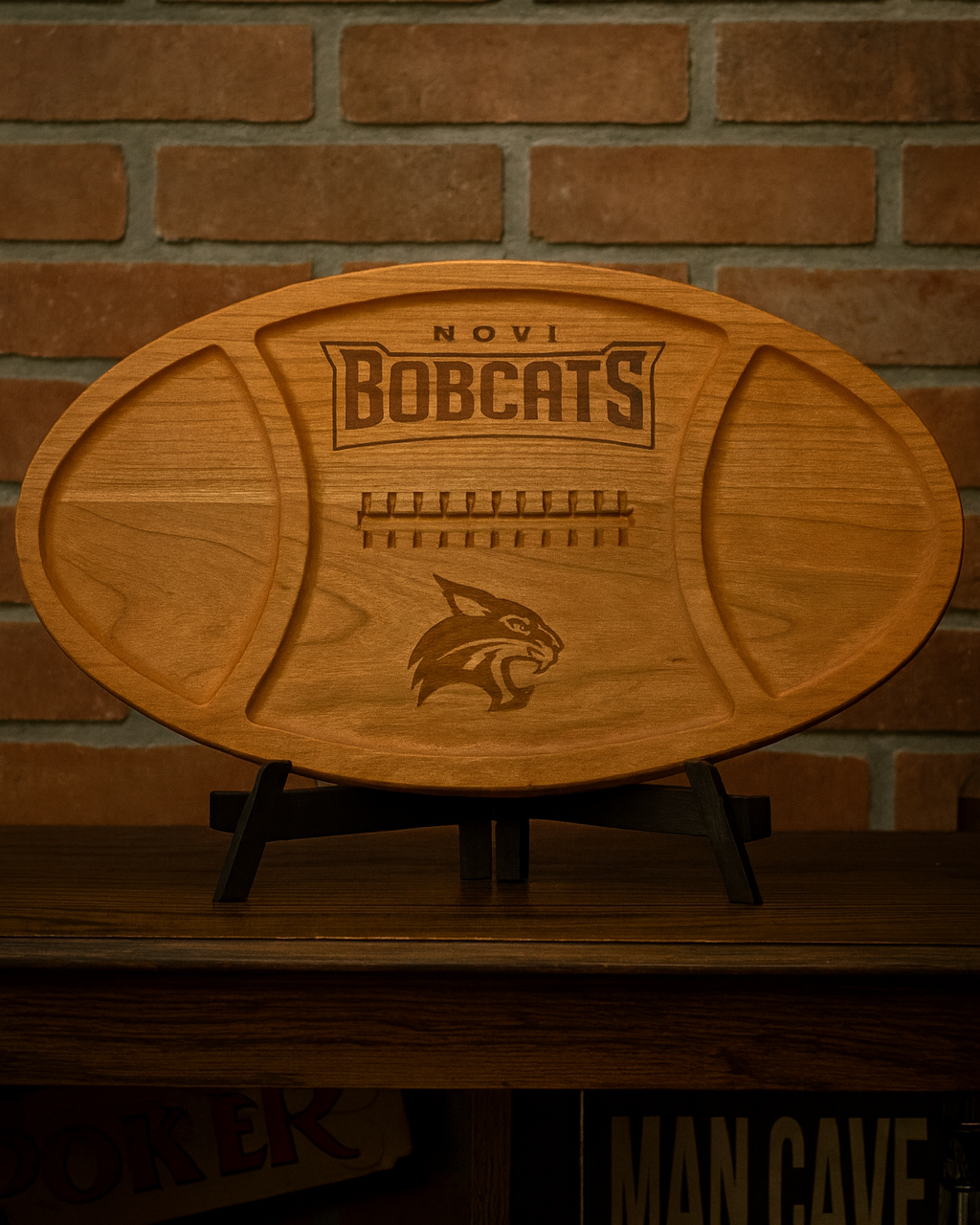 Wooden football-shaped cutting board with 'Novi Bobcats' engraving on a brick wall background.