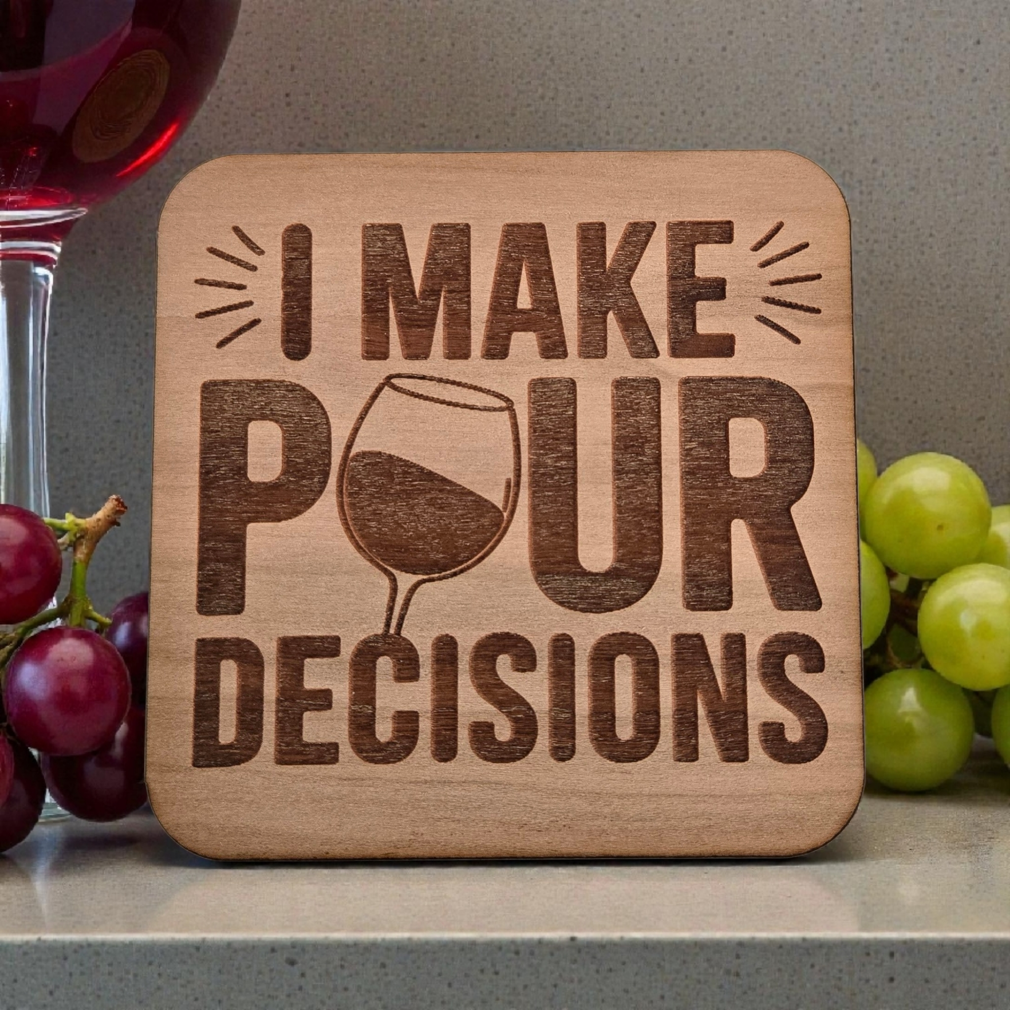 Wooden coaster with 'I Make Pour Decisions' text and a wine glass graphic, surrounded by grapes and a glass of red wine.
