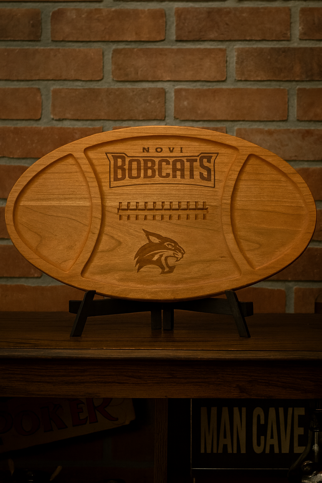 Wooden football-shaped cutting board with 'Novi Bobcats' engraving on a brick wall background.